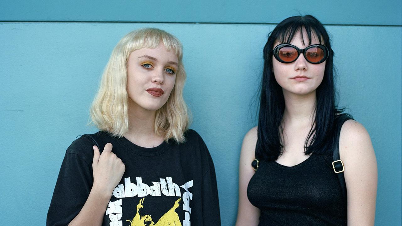 Zwei junge Frauen/Teenager-Mädchen stehen vor einer blauen Wand und blicken in die Kamera. Die linke trägt ein Black-Sabbath-T-Shirt, die rechte eine große Sonnenbrille mit orangefarbenen Gläsern. Zwei junge Frauen/Teenager-Mädchen stehen vor einer blauen Wand und blicken in die Kamera. Die linke trägt ein Black-Sabbath-T-Shirt, die rechte eine große Sonnenbrille mit orangefarbenen Gläsern.
