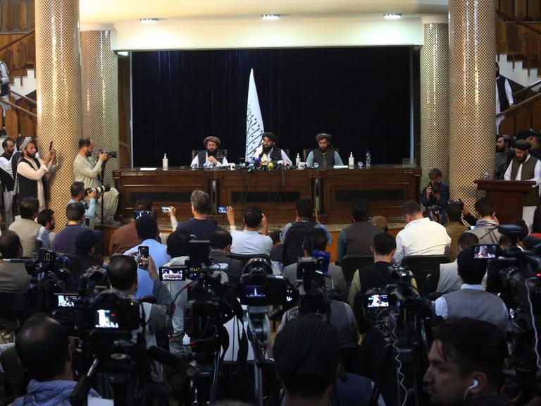 Der Taliban-Sprecher Zabihullah Mujahid sitzt mit zwei weiteren Männern am 17.08.2021 in Kabul auf einer Bühne, der Raum davor ist gefüllt mit Presseleute, die die Konferenz filmen und fotografieren.