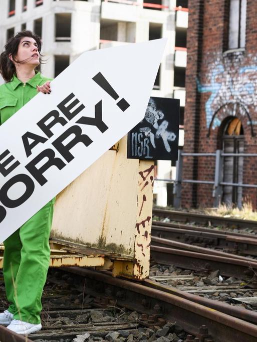 Alicia Agustín steht auf Bahngleisen und hält ein Schild mit der Aufschrift "We Are Sorry!"; im Hintergrund eine Baustelle.
