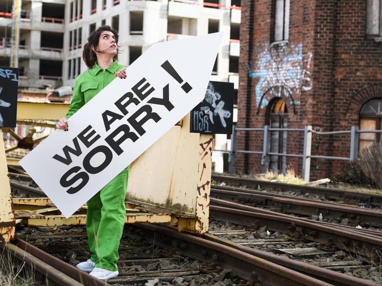 Alicia Agustín steht auf Bahngleisen und hält ein Schild mit der Aufschrift "We Are Sorry!"; im Hintergrund eine Baustelle.
