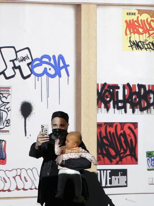 Ein Mann mit Maske und Baby im Arm fotografiert sich mit seinem Handy vor einem Bildschirm, auf dem die Rückseite eines Bilderrahmens zu sehen ist.