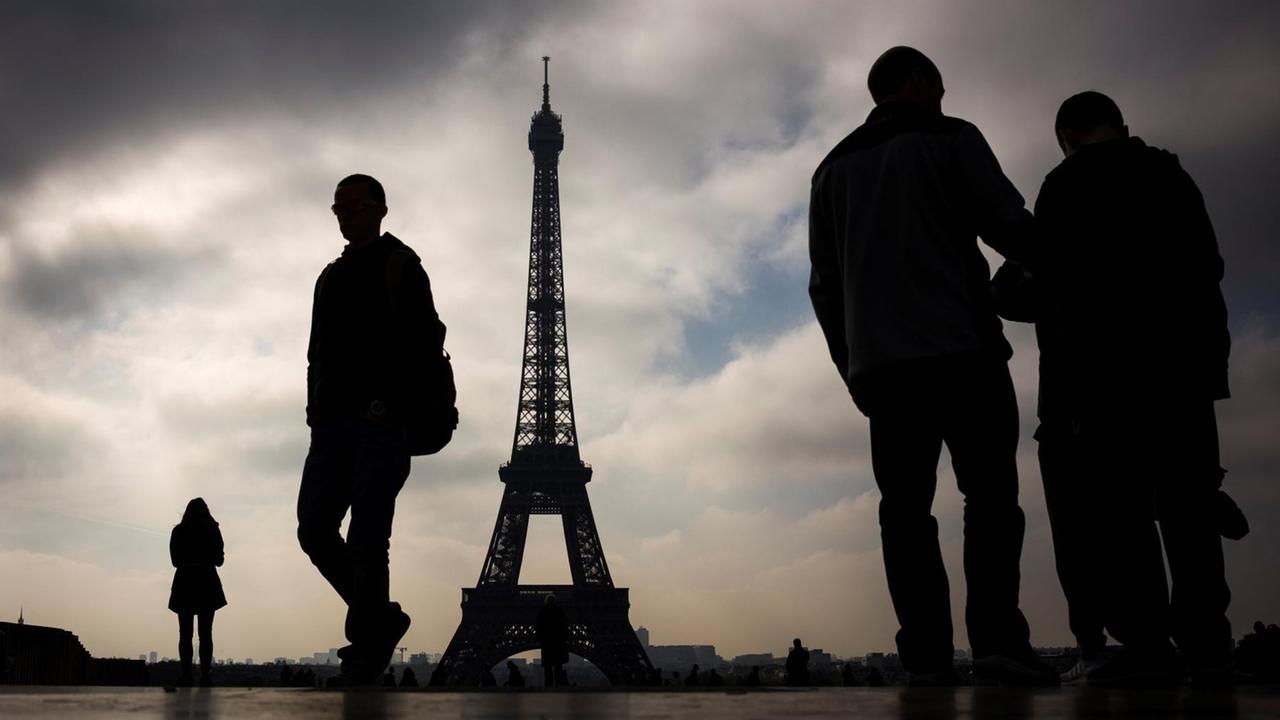 Eiffelturm in Paris, im Vordergrund stehen Menschen im Gegenlicht, der Himmel ist wolkenverhangen.