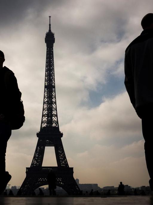 Das Bild zeigt den Eiffelturm in Paris, im Vordergrund stehen Menschen im Gegenlicht, der Himmel ist wolkenverhangen.