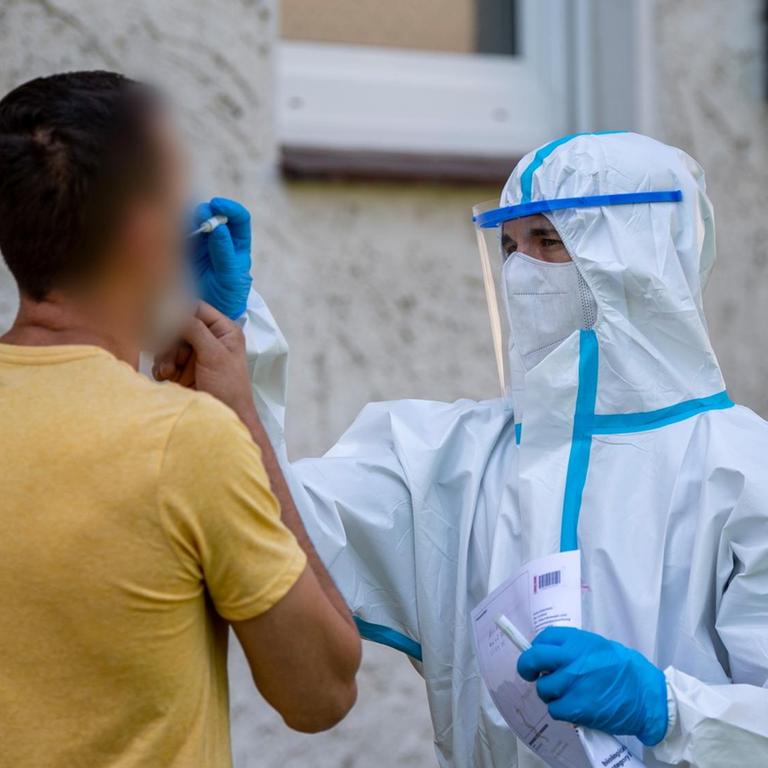 Ein Helfer vom Deutschen Roten Kreuz (DRK) nimmt vor einem Haus bei einem Mann einen Abstrich vor. 40 mobile Testteams sind unterwegs, um Angestellte der Firma Tönnies zu Hause in ihrer Quarantäne aufzusuchen. Sie überprüfen, ob diese dort anzutreffen sind und testen auch die Menschen, die mit ihnen in einem Haushalt leben. 