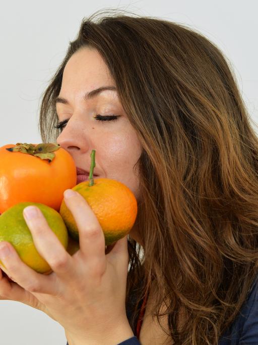 Eine junge Frau hält Äpfel, Mandarinen und eine Kaki in den Händen und riecht daran.