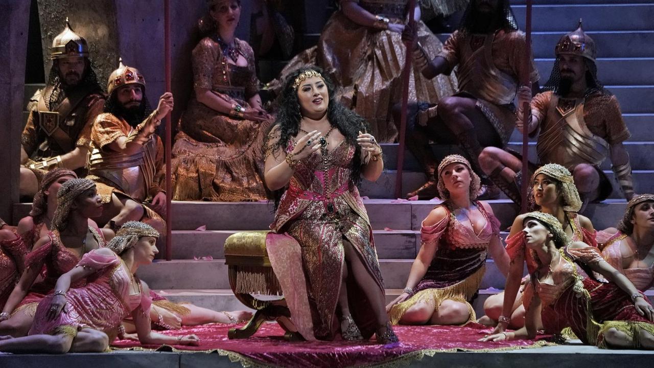 Dalila sitzt von Tänzerinnen umringt in einem orientalischen Gewand und hofft auf Erfolg bei Samson.