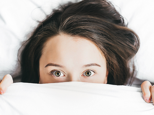 Eine Frau liegt im Bett und zieht angsterfüllt die Bettdecke bis unter die Augen.