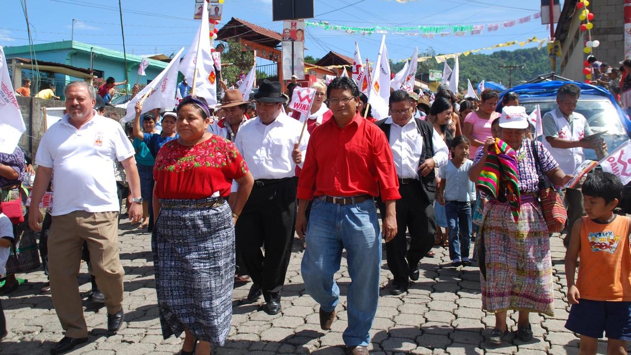   14 Millionen Guatemalteken sind 2011 aufgerufen ihrennächsten Präsidenten zu wählen - oder zum ersten Maleine Präsidentin?  Rigoberta Menchú: außerhalb des Landes die bekanntesteGuatemaltekin, Menschenrechsaktivistin undFriedennobelpreisträgerin von 1992. tritt  anfür die Frente amplio, die "Breite Front", ein losesWahlbündnis von vier linken Parteien.Tür zuschlagen, aus Auto steigen, im Hintergrund  Menchu anErz Die Kandidatin hält Einzug in San Pedro Yepocapa, einemAckerbürgerstädtchen von 8.000 Einwohnern, Mayas vomVolk der Kakchiquel, die meisten traditionell gekleidet;der Pazifik ist in Sichtweite, Vulkan Fuego, "Feuer",thront über dem Ort. Gerade hat er unter FauchenRauchkringel in den Himmel geschickt. Die vielleicht 300Menschen auf dem Marktplatz nehmen keine Notizdavon. Pablo Monsanto