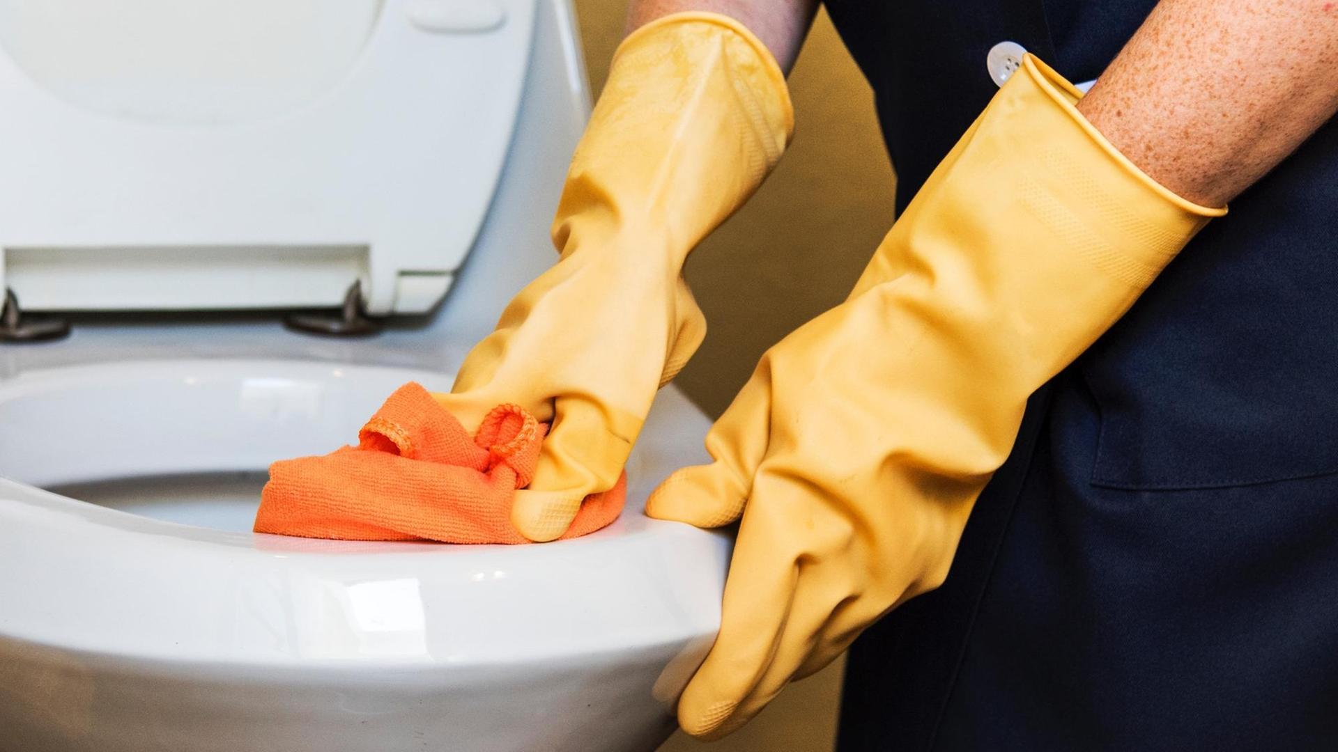 Hände in gelben Handschuhen putzen mit einem orangenen Tuch eine Toillette
