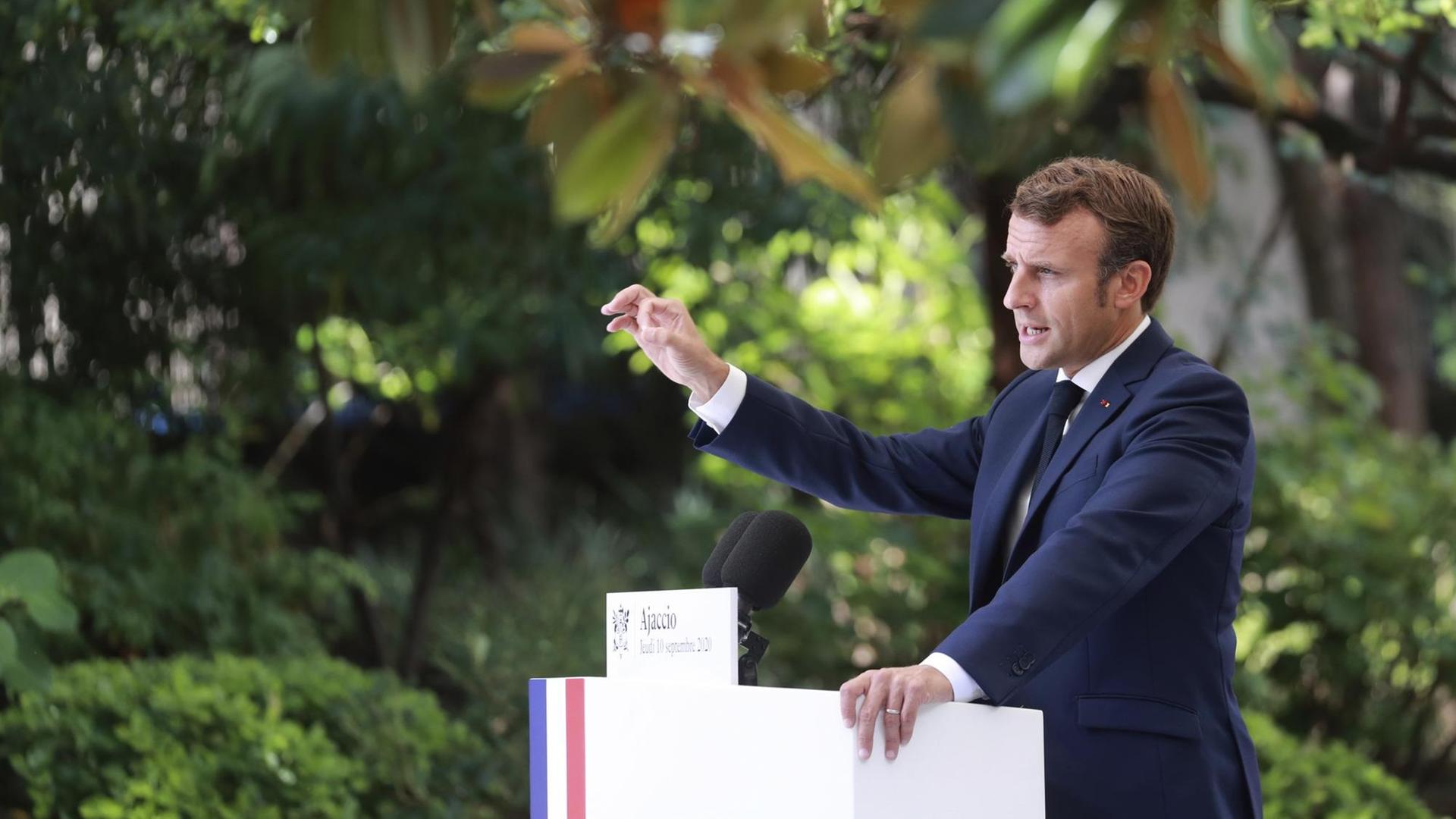 Macron steht unter Bäumen an einem Rednerpult mit den französischen Farben, spricht in ein Mikrofon und gestikuliert mit der erhobenen Hand. Er ist von der Seite fotografiert.