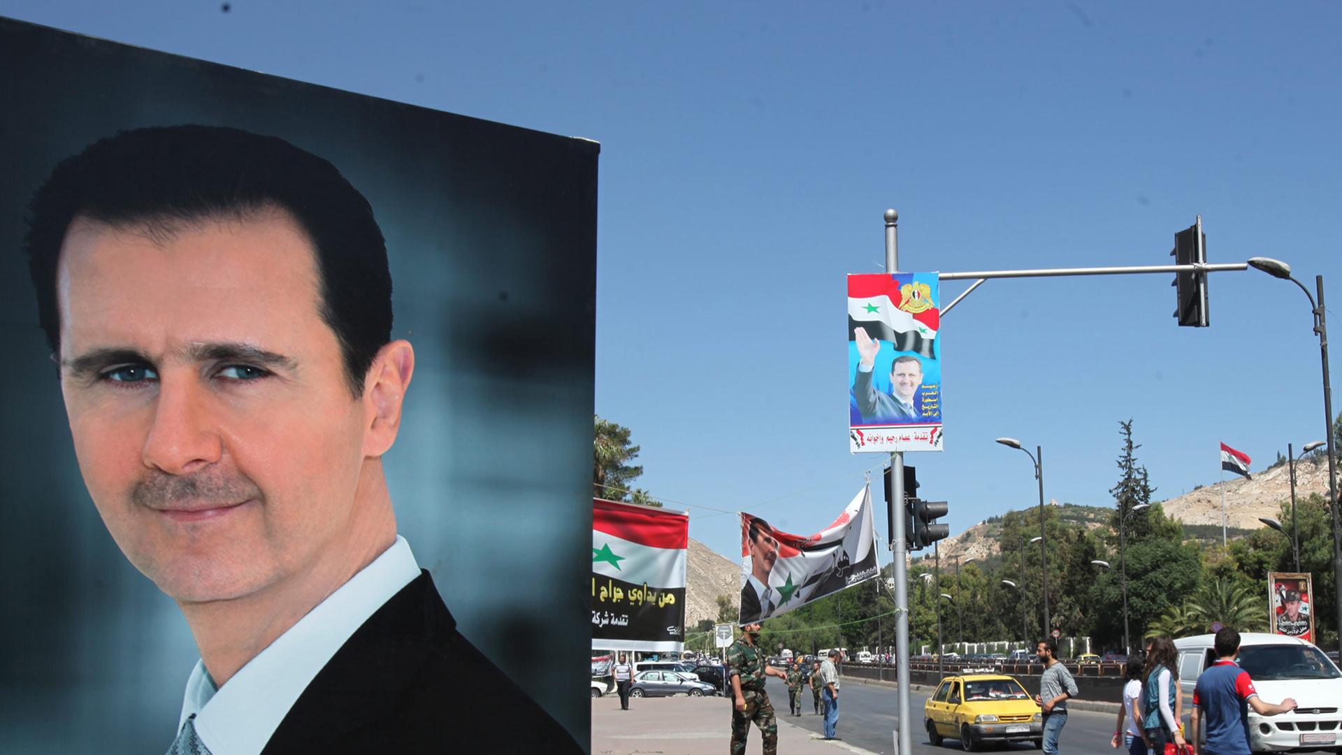 Großes Portrait von Assad auf einem Wahlplakat an einer Straße, im Hintergrund Straßenszene mit Autos und Fußgängern, an einer Straßenlampe ein kleineres Wahlplakat eines Oppositionellen.