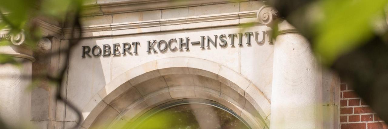  Der Eingang des Robert Koch-Instituts ist mit seinem Namenslogo durch Bäume hindurch zu sehen. 