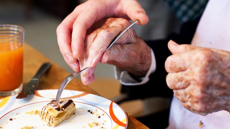 Zu sehen sind die Hände einer älteren Frau beim Essen. Eine andere Person legt ihre Hand auf die Hand der Frau. Das zeigt Unterstützung, Betreuung und Fürsorge. Zu sehen sind die Hände einer älteren Frau beim Essen. Eine andere Person legt ihre Hand auf die Hand der Frau. Das zeigt Unterstützung, Betreuung und Fürsorge.