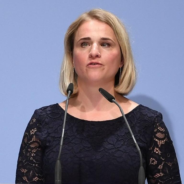 Eine blonde Frau steht an einem Rednerpult und spricht.