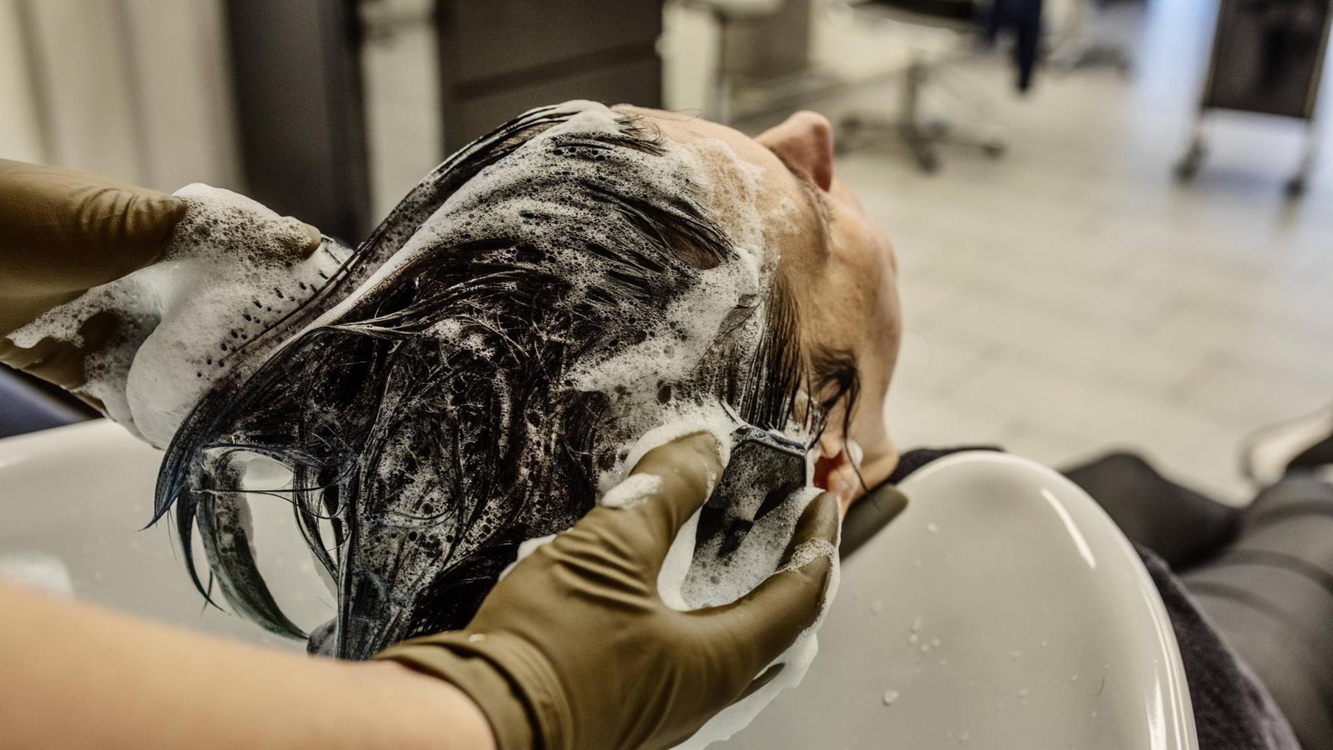 Eine Frau sitzt im Frisiersalon und hat den Kopf ins Waschbecken zurückgelegt. Sie hat Schaum im Haar, die behandschuhten Hände des Friseurs sind zu sehen, wie sie der Frau Schaum aus dem Haar spülen.