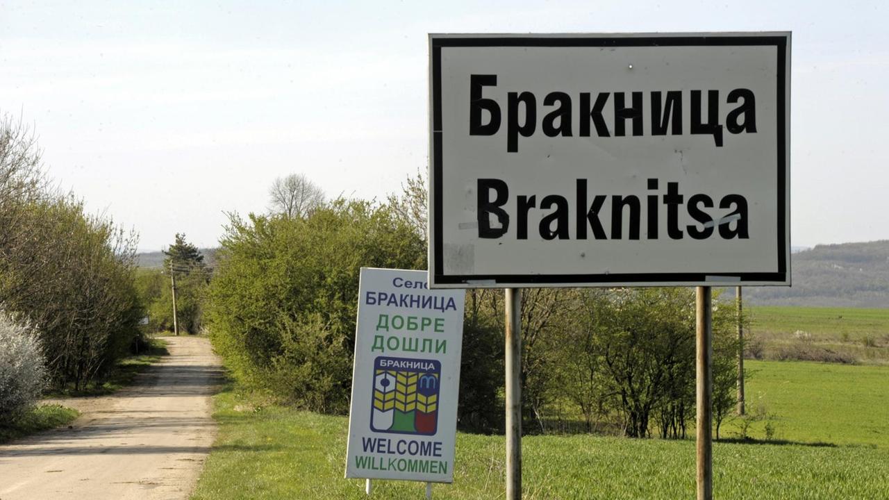 Eine Wiese mit einem Stadtschild, auf dem Braknitsa steht. Dahinter ein weiteres Schild mit Willkommensgrüßen in verschiedenen Sprachen.