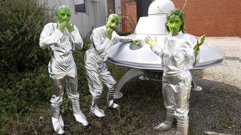 Drei als Außerirdische Verkleidete mit grüngeschminkten Gesichtern und aufgeklebten Fühlern stehen in silbernen A^nzügen vor einem UFO.