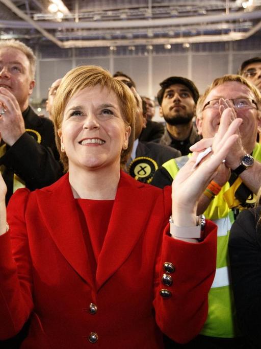 Sturgeon steht in einem roten Kostüm zwischen Parteifreunden, lächelt und klatscht.