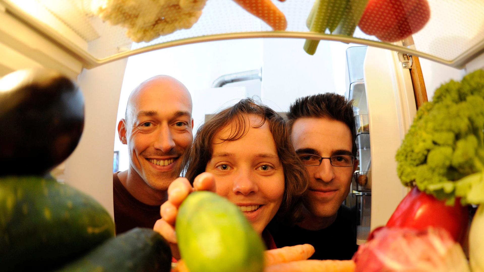 Bild aus einem Kühlschrank fotografiert: Drei junge Menschen drängen sich vor der Tür; eine Frau greift nach Gemüse im Kühlschrank.
