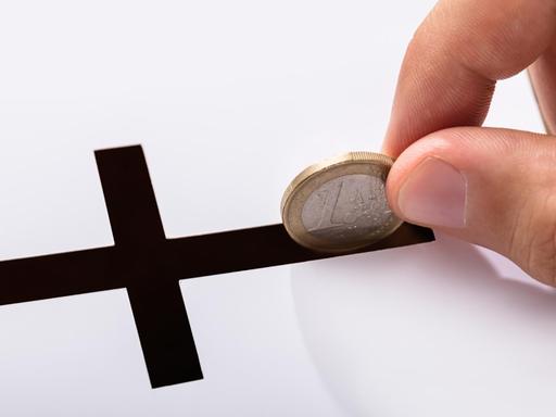 Eine Hand steckt einen Euro in einen schwarzen Schlitz in Kreuzform (Symbolbild).