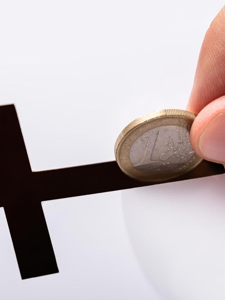 Eine Hand steckt einen Euro in einen schwarzen Schlitz in Kreuzform (Symbolbild).