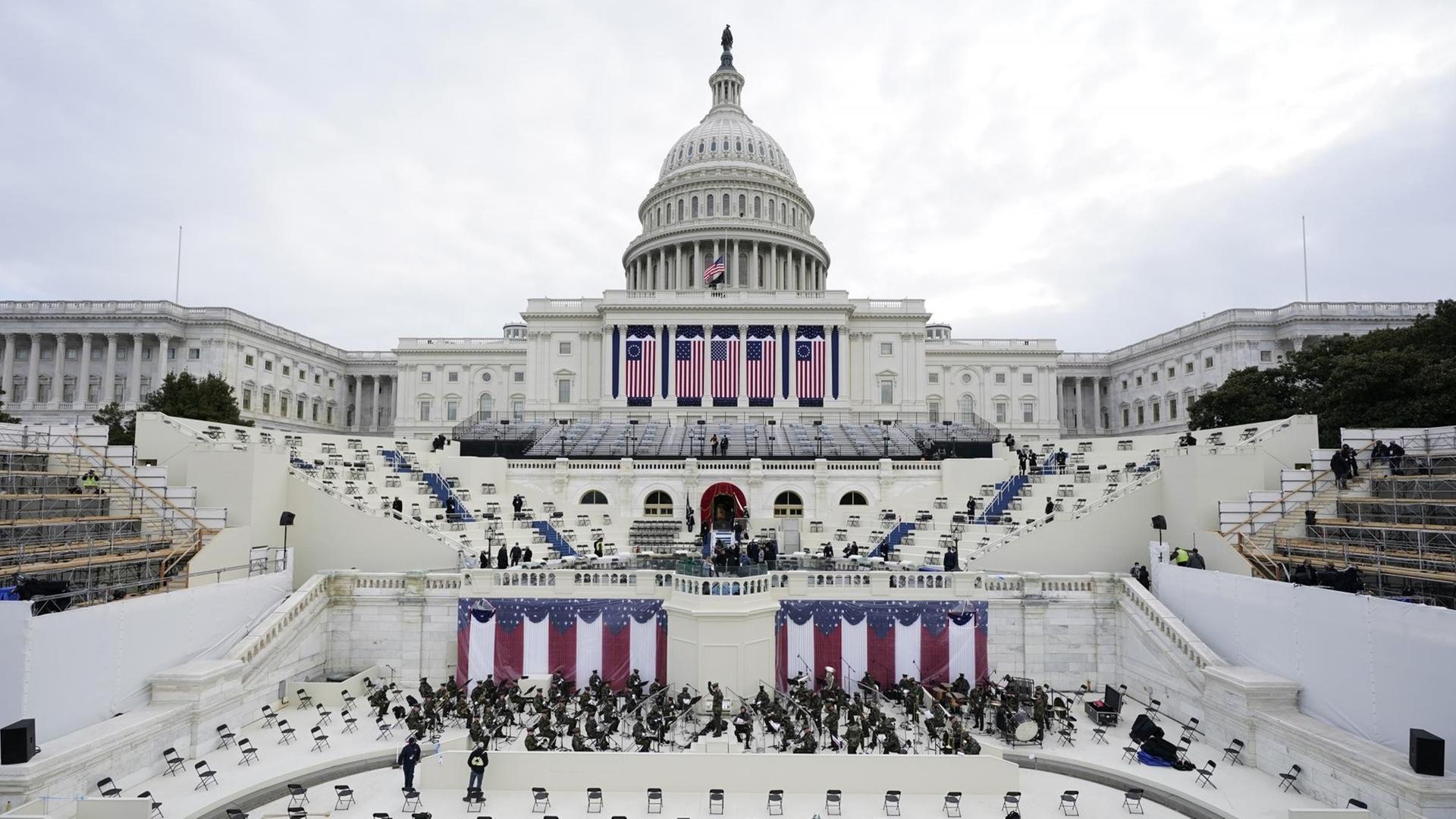 Blick auf das Kapitol in Washington mit herabhängenden großen US-Fahnen und einer probenden Militärkapelle, davor mit großem Abstand leere Stühle