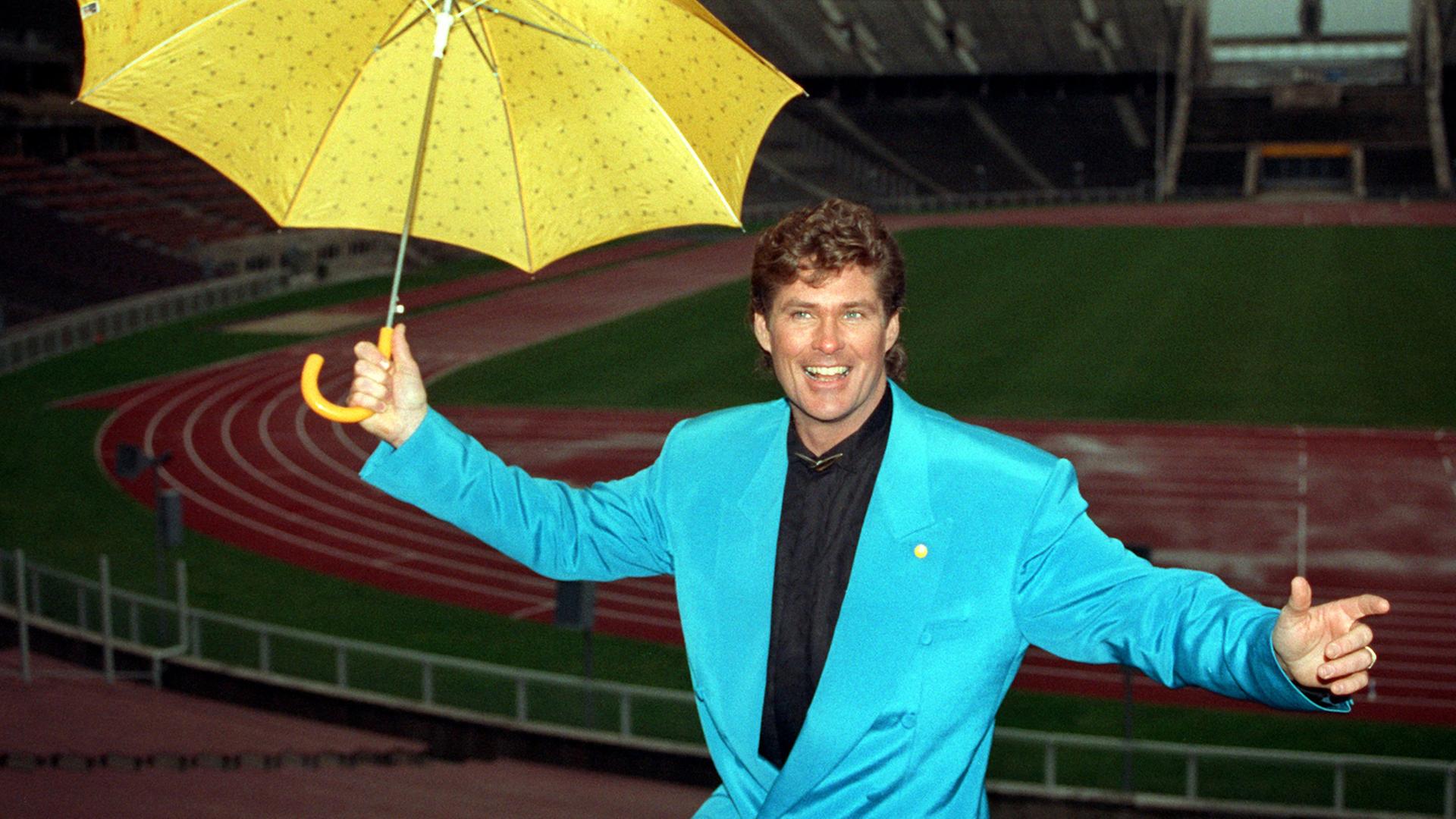 Der amerikanische Popstar David Hasselhoff präsentiert sich lächelnd mit einem gelben Regenschirm im türkisen Anzug.