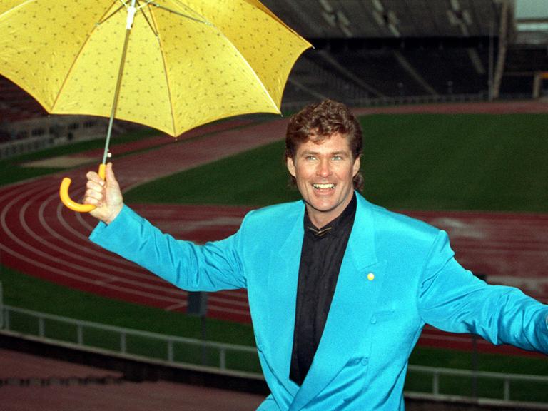 Der amerikanische Popstar David Hasselhoff präsentiert sich lächelnd mit einem gelben Regenschirm im türkisen Anzug.
