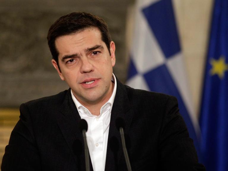 Alexis Tsipras bei einer Pressekonferenz an einem Rednerpult, im Hintergrund die griechische Fahne.