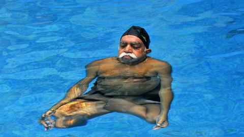 Ein indischer Mann mit weißem Schnurrbart praktiziert Yoga in einem Schwimmbecken.