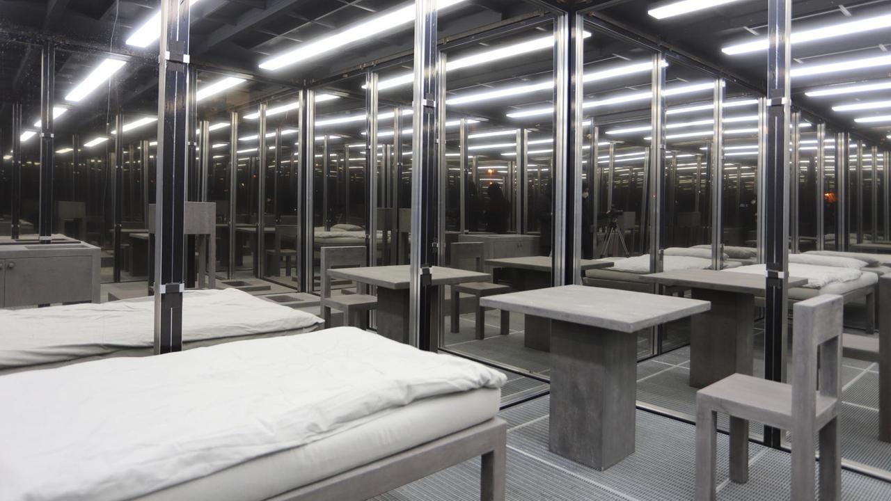 Installation einer freistehenden, transparenten Zelle, mit leeren Betten und grellen Neunleuchten.