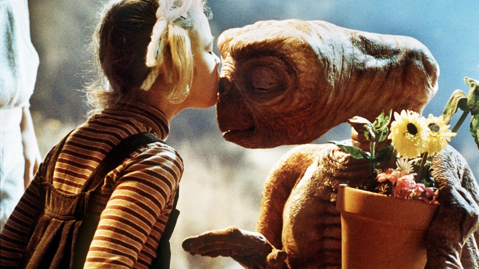 Ein junges, blondes Mädchen mit Latzhose und zwei Zöpfen küsst einen schrumpeligen Außerirdischen auf die Stirn. Er hält einen Blumentopf links vor sich und hat die Augen geschlossen.