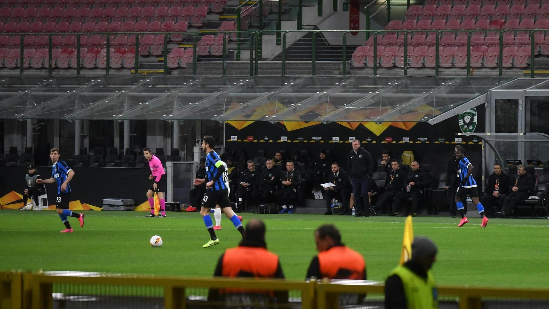 Speiele rvon Inter Mailand und Rasgrad in einer Spielszene vor leeren Zuschauerrängen. Im Vordergrund eine Bank mit zwei Männern in orangefabenen Sicherheitswesten.