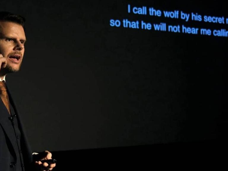 Der Künstler steht an einem Mikrophon, hinter ihm erscheinen blaue Textzeilen auf dem Bühnenhintergrund.