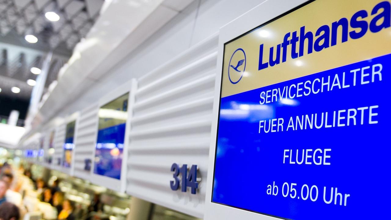 An einem Flugschalter der Lufthansa in Frankfurt am Main ist auf dem Display zu lesen: "Serviceschalter für annulierte Flüge ab 5 Uhr." Vor dem Schalter sind unscharf Fluggäste zu erkennen; dahinter sitzen zwei Service-Angestellte der Lufthansa.