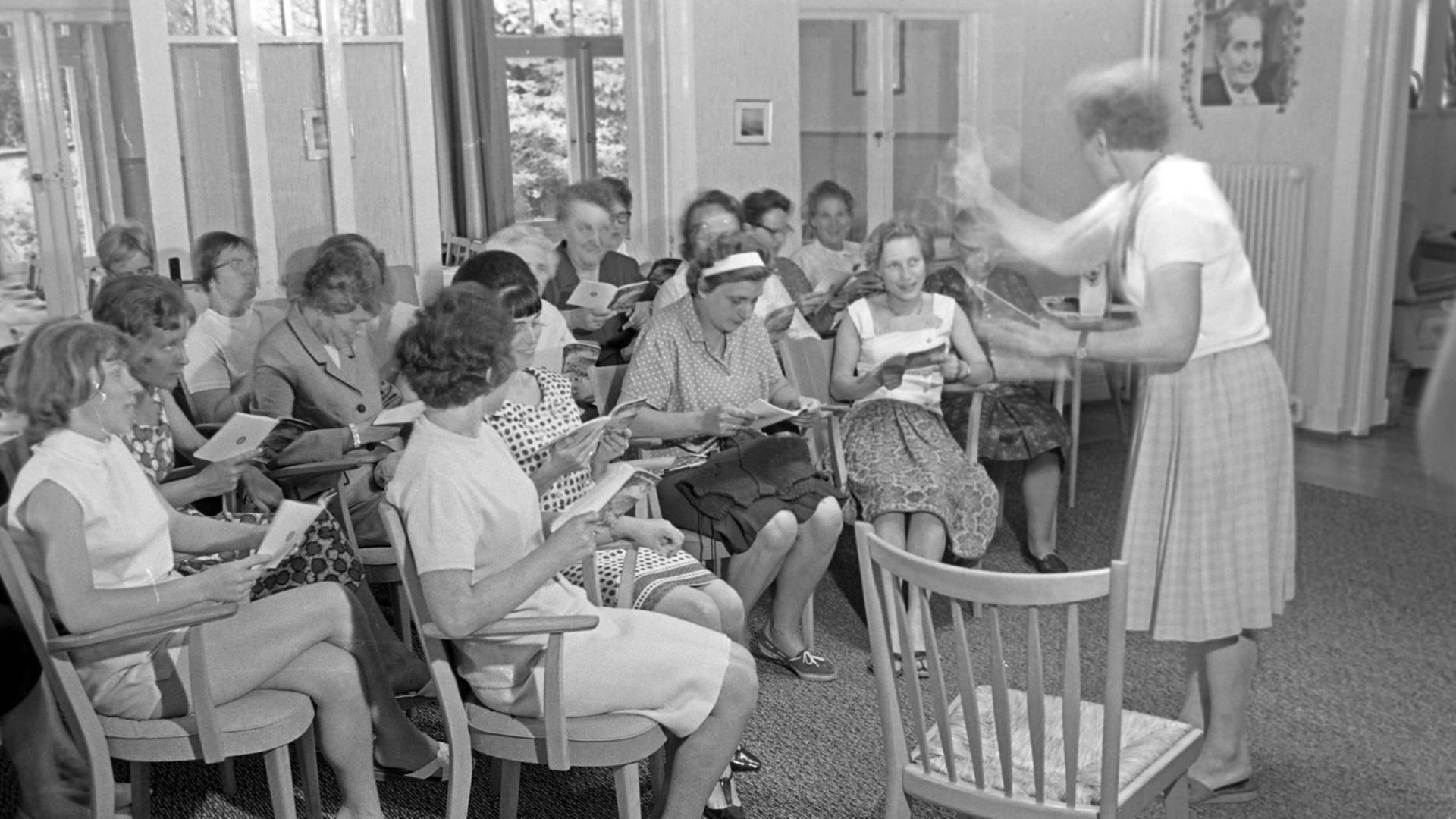 Das Foto ist schwarz-weiß. Frauen sitzen in einem Raum auf Stühlen im Halbkreis und singen, rechts steht eine weitere Frau und dirigiert. Im Hintergrund an der Wand rechts hängt ein Porträt von Elly Heuss-Knapp.