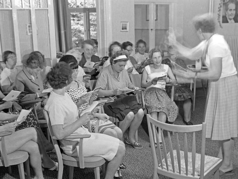 Das Foto ist schwarz-weiß. Frauen sitzen in einem Raum auf Stühlen im Halbkreis und singen, rechts steht eine weitere Frau und dirigiert. Im Hintergrund an der Wand rechts hängt ein Porträt von Elly Heuss-Knapp.