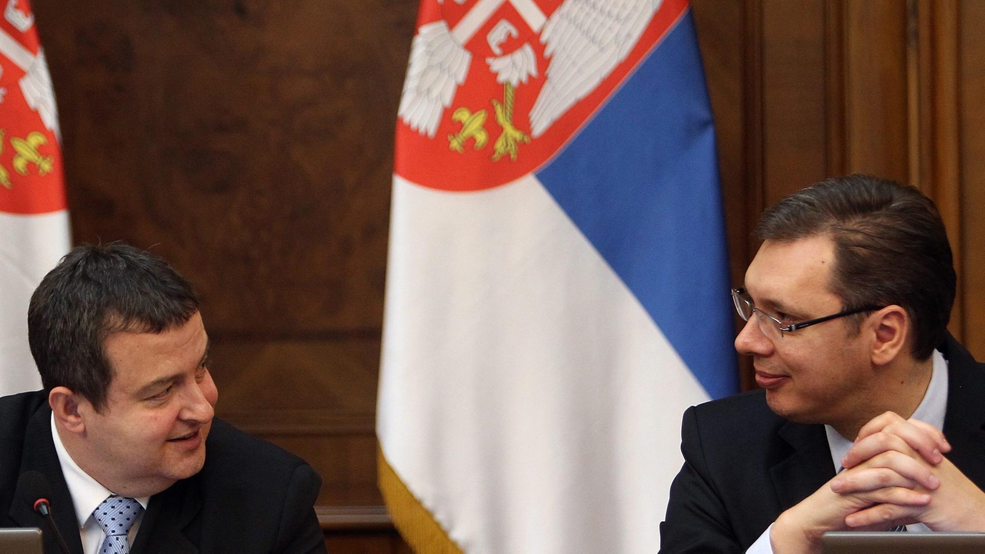 Ivica Dacic und Alexander Vucic sitzen an einem Tisch vor Laptops und unterhalten sich. Im Hintergrund vor einer branuen Holzwand hängen zwei serbische Flaggen in den Farben blau-weiß-rot.