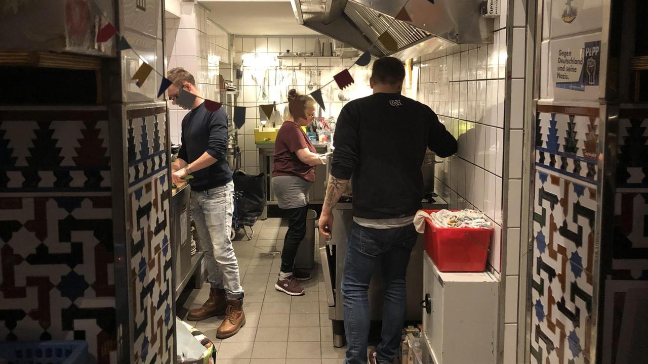 Eine enge Küche, oben begrenzt durch eine große Abzugshaube, stehen drei Menschen und arbeiten.