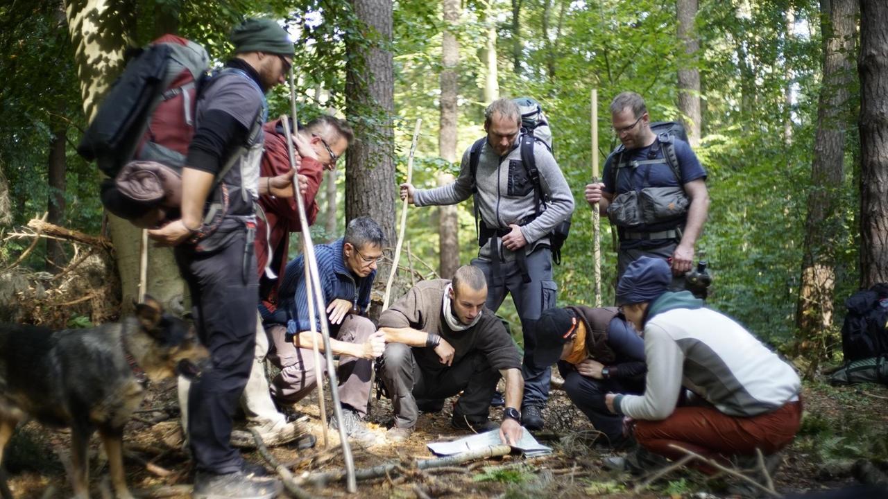 Eine Gruppe von Menschen in Outdoorkleidung und mit Outdoorausrüstung steht im Wald und schaut gemeinsam auf eine Karte.