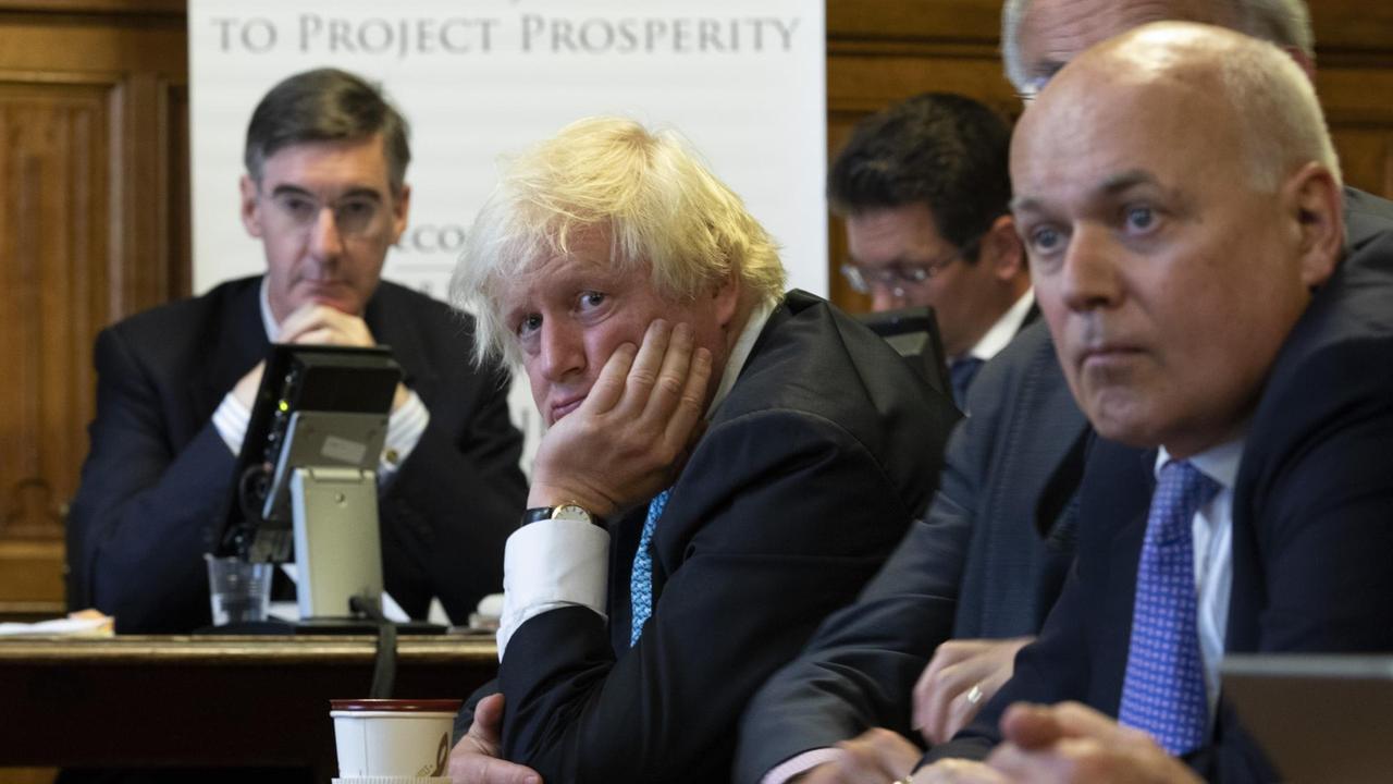 Boris Johnson stützt seinen Kopf auf die Hand und schaut müde an der Kamera vorbei. Um ihn herum unterschiedliche Personen, u.a. Jacob Rees Mogg.