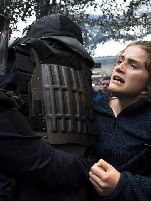 Eine junge Frau wird von mehreren vermummten Polizisten bedrängt und festgehalten. Sie hat ein ernstes Gesicht.