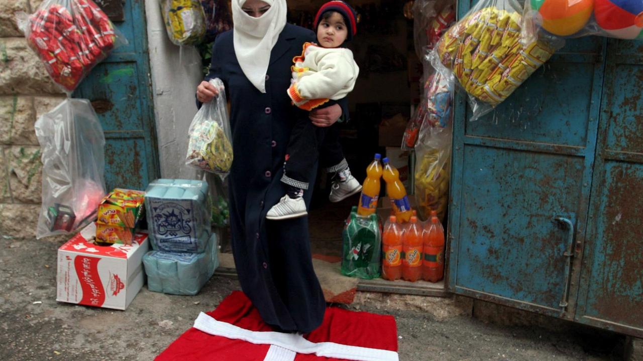 Eine Frau in Hebron trägt ihr Kind und ihre Einkäufe über eine dänische Fahne.