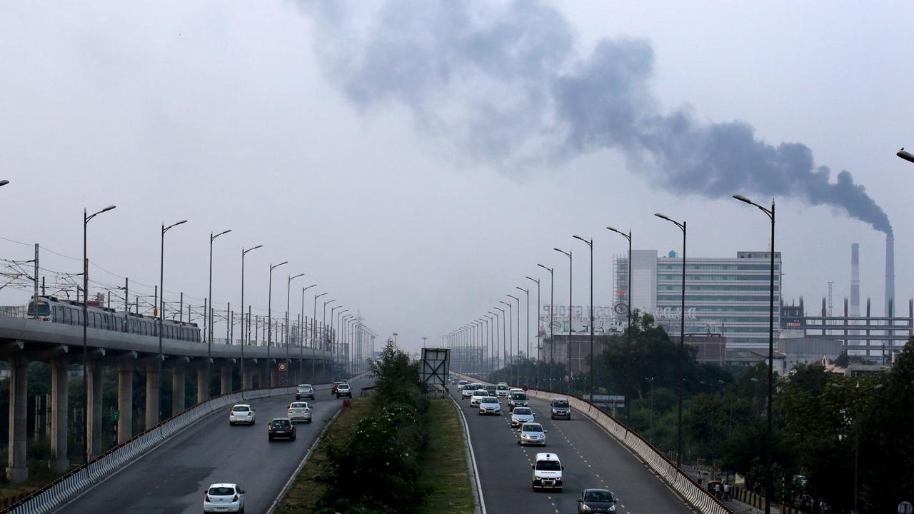 Eine stark befahrene Schnellstraße in Neu Delhi in Indien führt an einer Fabrik mit rauchendem Schlot vorbei. Das Bild ist blaustichig, der Himmel grau. Das Foto wurde wohl von einer Brücke aus aufgenommen.