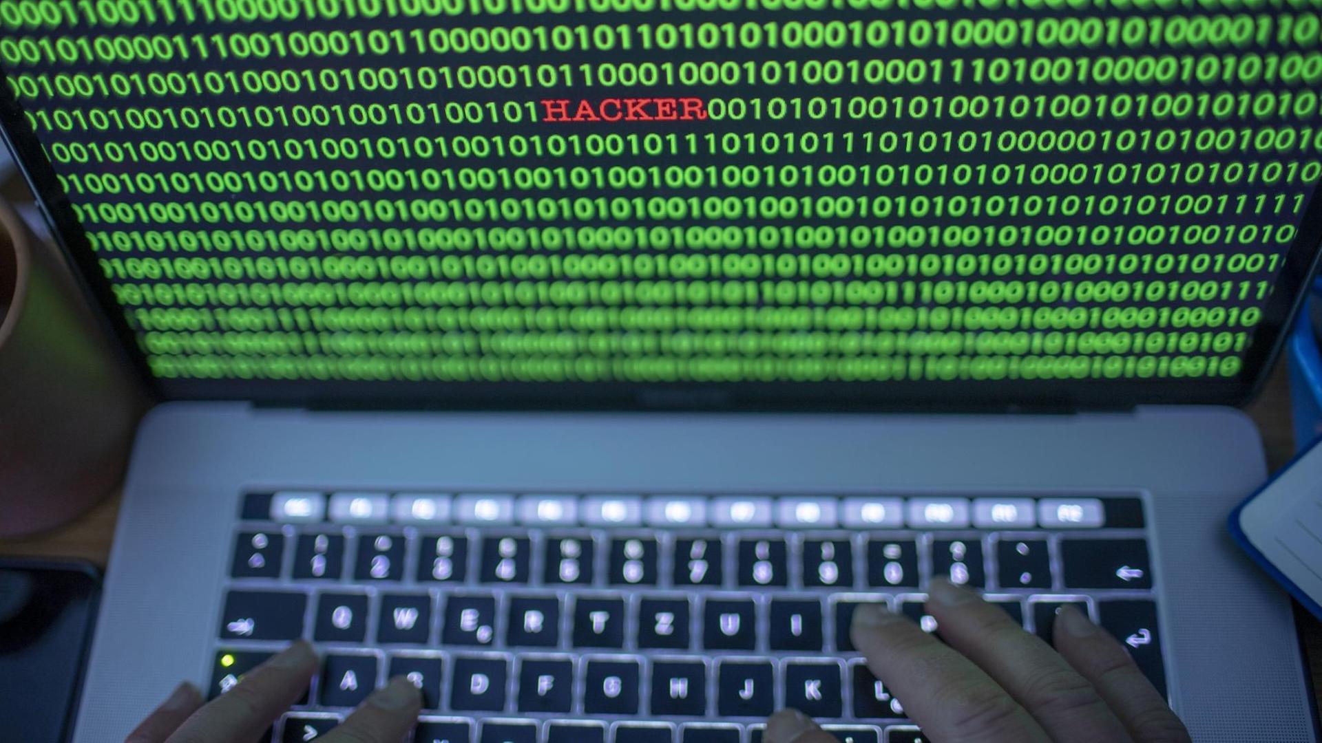 Zwischen dem Binärcode auf einem Laptopmonitor ist der Schriftzug "Hacker" zu sehen