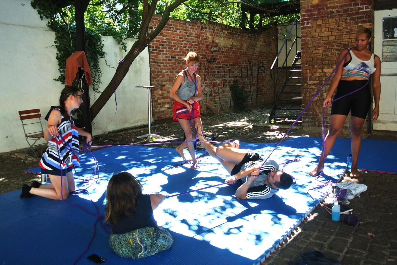 Das Performancekollektiv Polymora Inc bei der Probe unter freiem Himmel. 5 Personen halten sich an einem Seil fest und sind somit alle miteinander verbunden. Die Person in der Mitte liegt auf dem Boden, als wäre sie die Beute der anderen.
