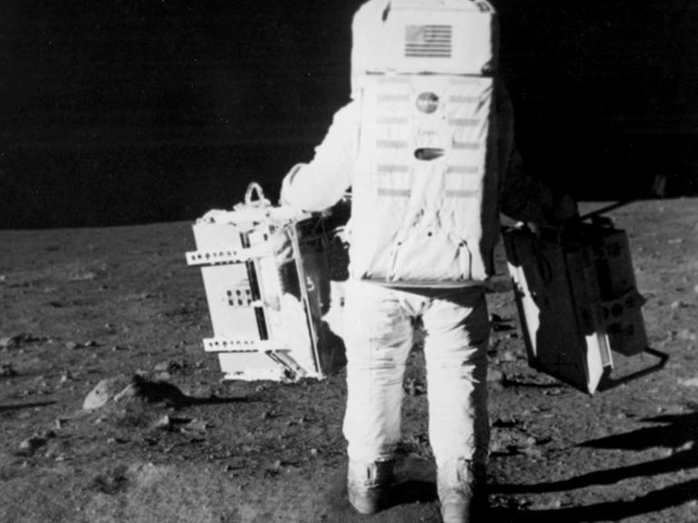 Der Astronaut Edwin E. Aldrin Jr. läuft über die Mondoberfläche und trägt zwei Taschen mit wissenschaftlichem Equipment bei sich.