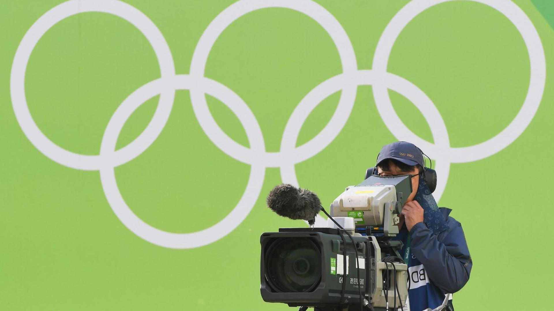 Ein TV-Kameramann filmt bei den Olympischen Spielen 2016 in Rio, hinter ihm sind die Olympischen Ringe zu sehen.