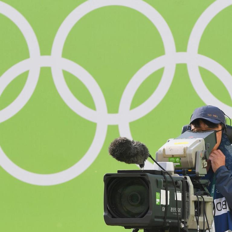 Ein TV-Kameramann filmt bei den Olympischen Spielen 2016 in Rio, hinter ihm sind die Olympischen Ringe zu sehen.