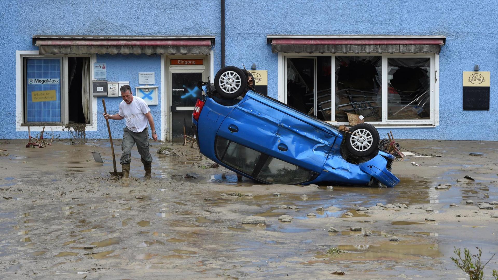 Ein Einwohner des Ortes Simbach am Inn in Bayern steht im Hochwasser-Gebiet auf einer überfluteten Straße neben einem beschädigten Auto, das auf dem Dach liegt.
